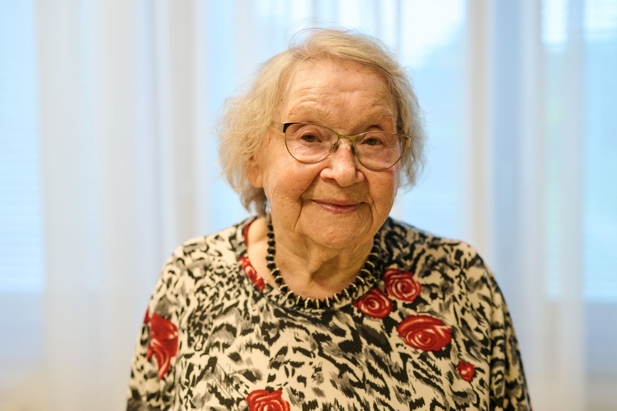 Eine Seniorin mit gemustertem Kleid schaut frontal in die Kamera.