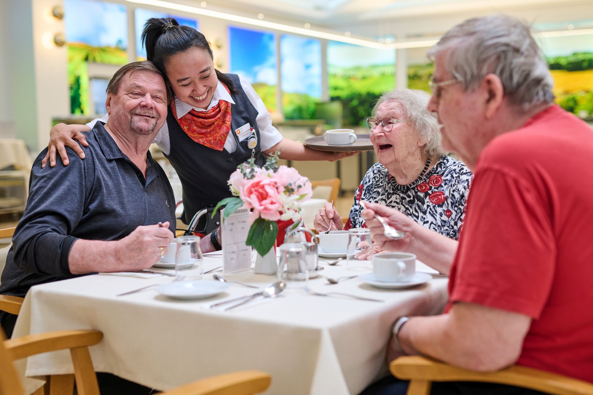 Eine ältere Frau und zwei ältere Herren sitzen lachend an einem Tisch und essen Suppe. Eine Servicekraft umarmt einen der Herren.