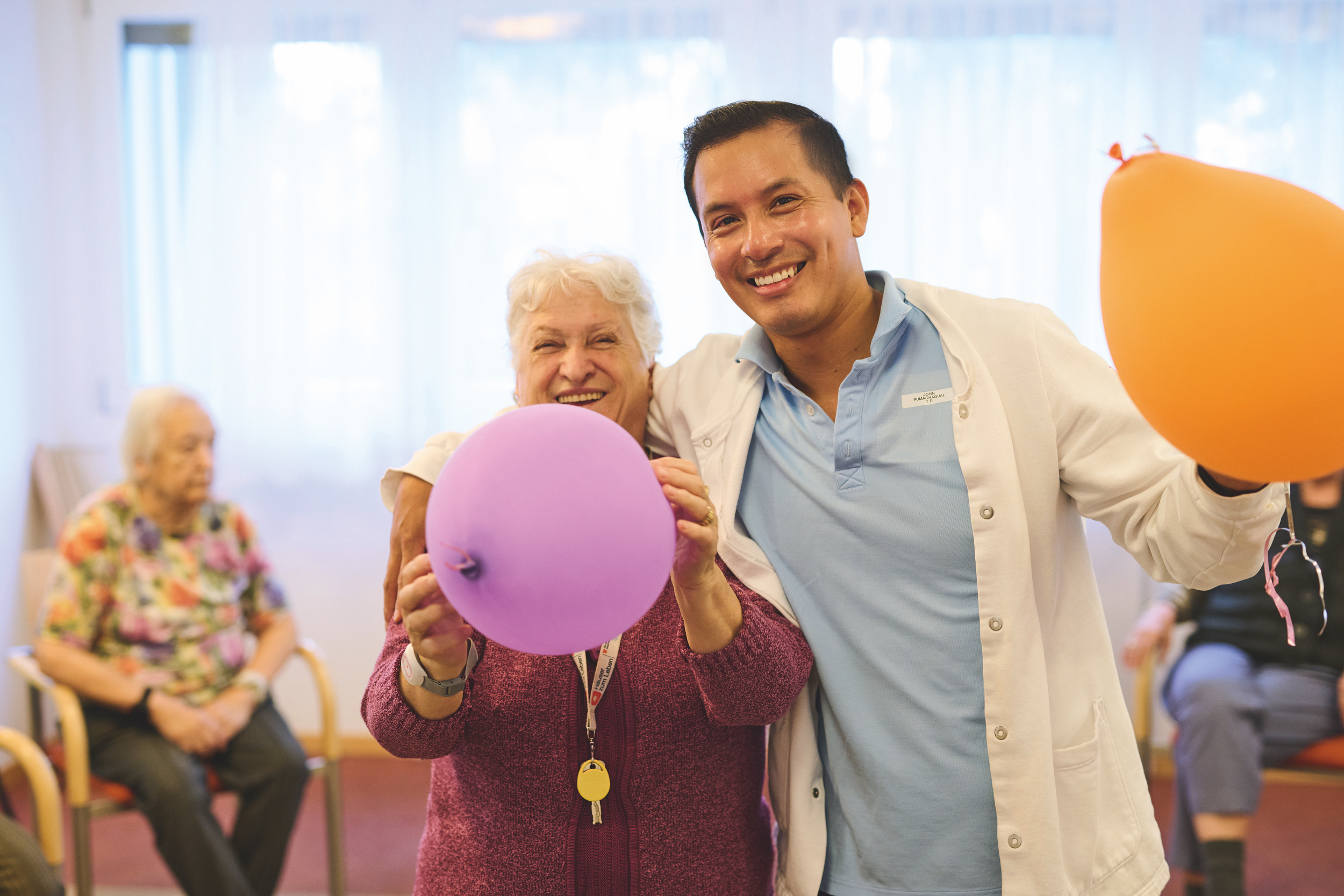 Ein Pfleger umarmt eine Seniorin. Beide halten einen Luftballon.